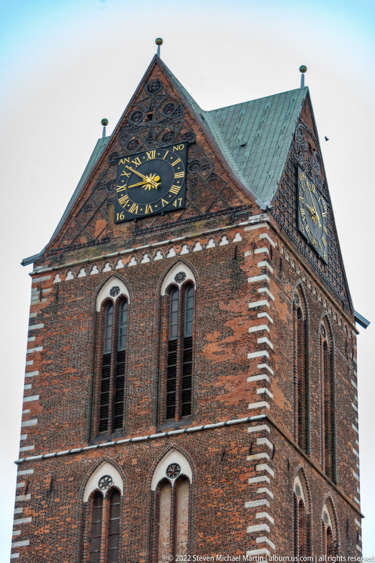 Upper brick tower with paired arched openings, clock faces, steep roof, and small finials.