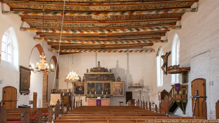 Whitewashed church nave with painted timber ceiling, chandeliers, pews, ornate pulpit, and large altarpiece at the far end.