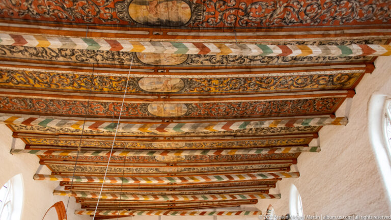 Close upward view of richly painted wooden ceiling beams with floral, geometric, and figurative ornament.