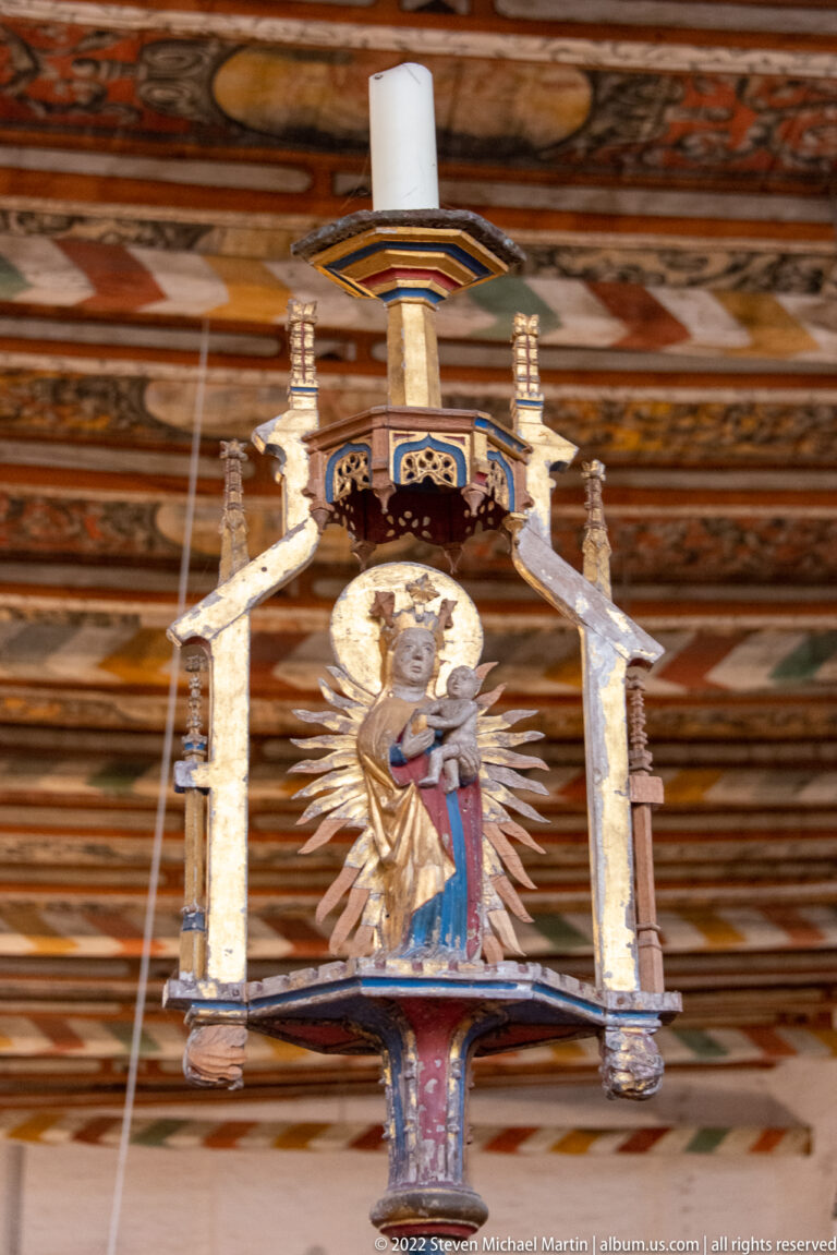 Polychrome devotional figure beneath a small canopy, backed by radiating rays and suspended below a painted wooden ceiling.
