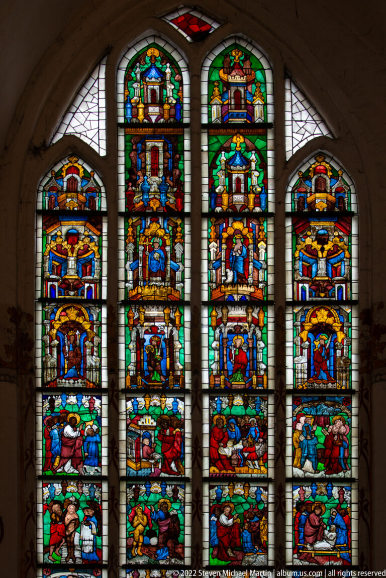 Large five-part stained-glass window filled with many colorful narrative scenes arranged in stacked registers.