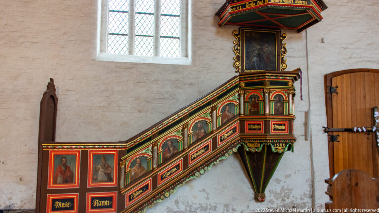 Polychrome wooden pulpit with painted stair panels, gilded trim, and polygonal sounding board mounted against a white wall.
