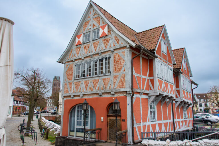 Orange half-timbered building with white framing, steep tiled roof, and decorative gable beside a canal.