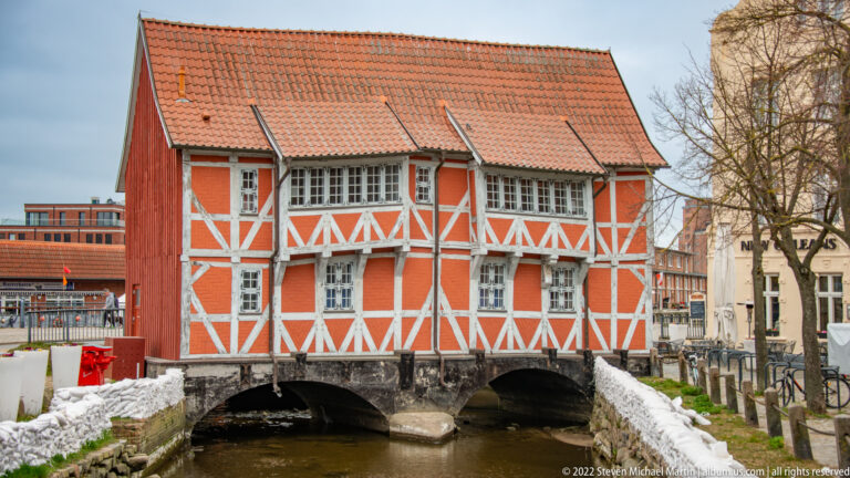 Orange-and-white half-timbered building spanning a narrow canal on masonry arches beneath a red-tiled roof.