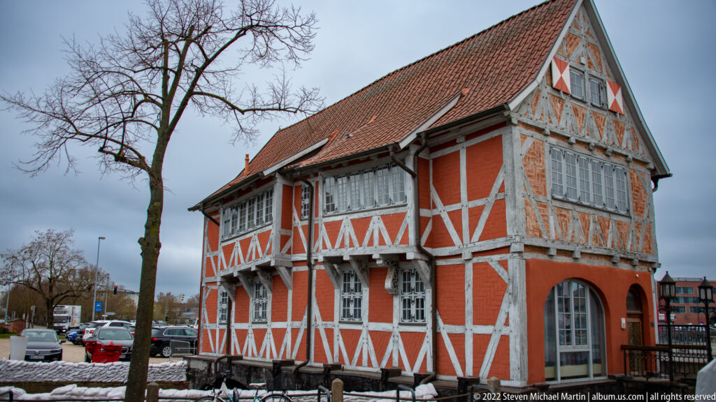 Oblique view of orange-and-white half-timbered canal building with steep roof, projecting upper story, snow, and nearby parked cars.