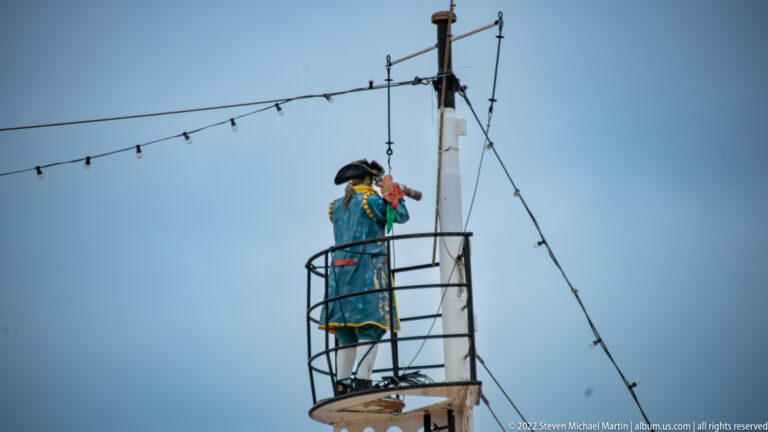 Costumed sailor figure in blue stands on a circular mast platform amid rigging and strings of lights against the sky.