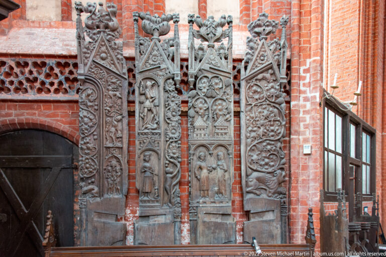 Four tall carved wooden Gothic panels with saints, tracery, foliage, and pointed architectural forms against a brick wall.