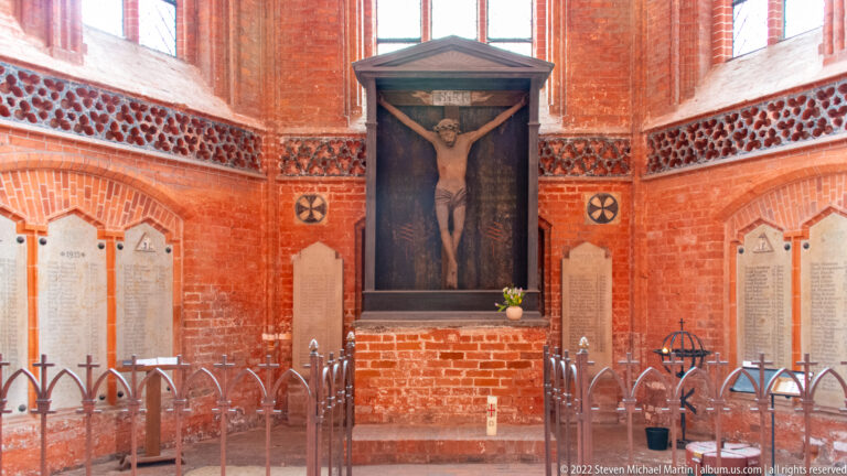 Large crucifix on a red-brick chapel wall between memorial tablets, with low wrought-iron rails in front.