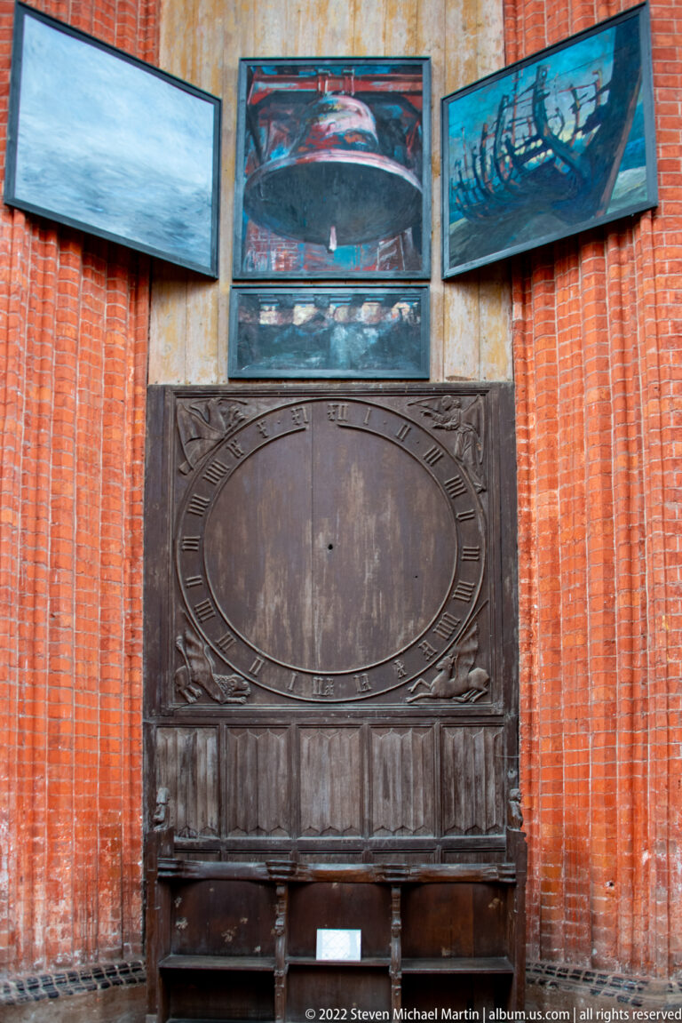Dark carved wooden chest or bench with an oval central panel stands below three modern paintings on a brick wall.