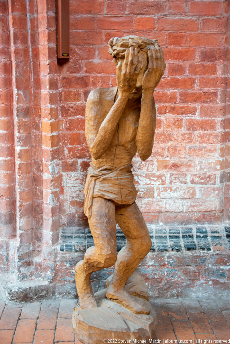 Carved wooden male figure crouches under a heavy load held above his shoulders against a red-brick wall.