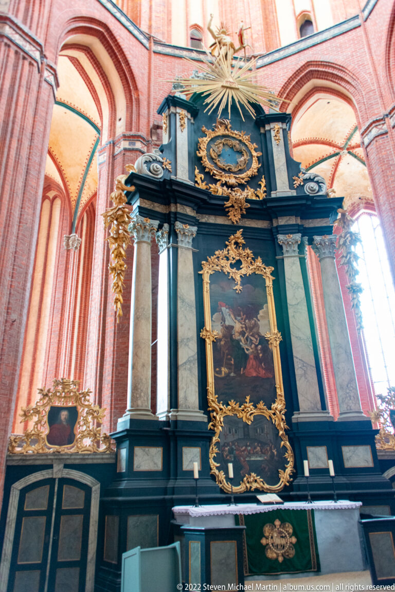 Large black-and-gold altar with columns, gilded scrollwork, central religious painting, and starburst ornament in a brick church.