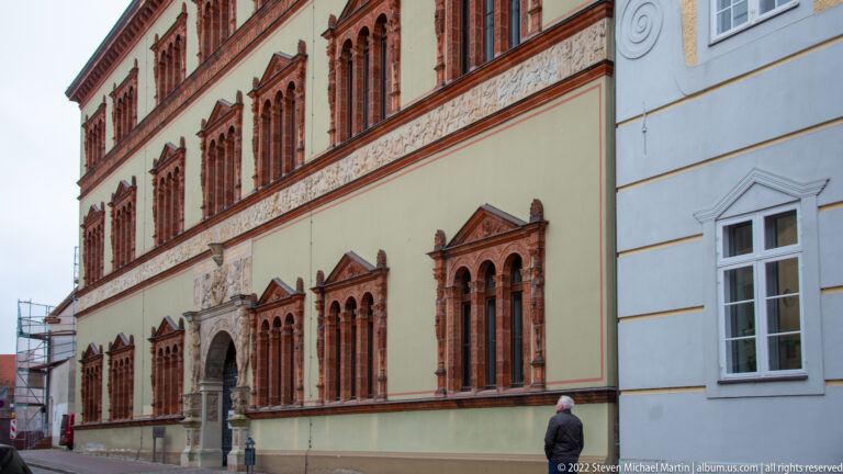 Long pale Renaissance façade with red-framed windows, decorated bands, and an ornate central portal.