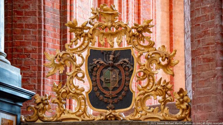 Gilded carved acanthus frame surrounds a dark heraldic shield mounted against a red-brick church wall.