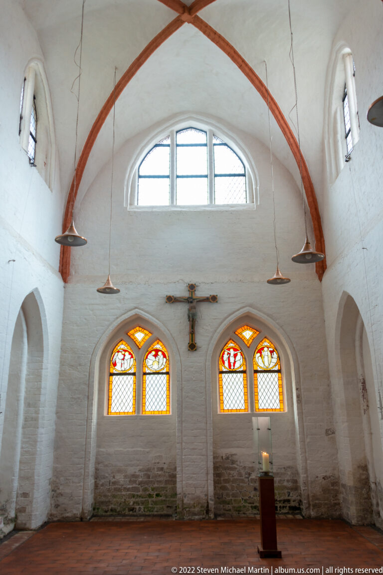Plain whitewashed vaulted church interior with red ribs, paired stained-glass windows, hanging lamps, and a wall crucifix.