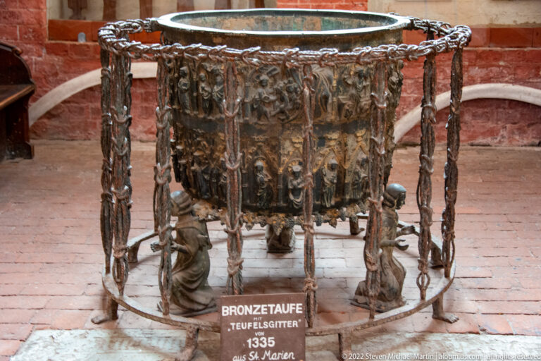 Large medieval bronze baptismal font with relief figures enclosed by an openwork chained metal frame; label gives the date 1335.