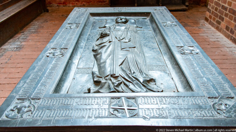 Bronze grave slab with relief of a praying robed cleric, border inscription, small heraldic shield, and corner figures.