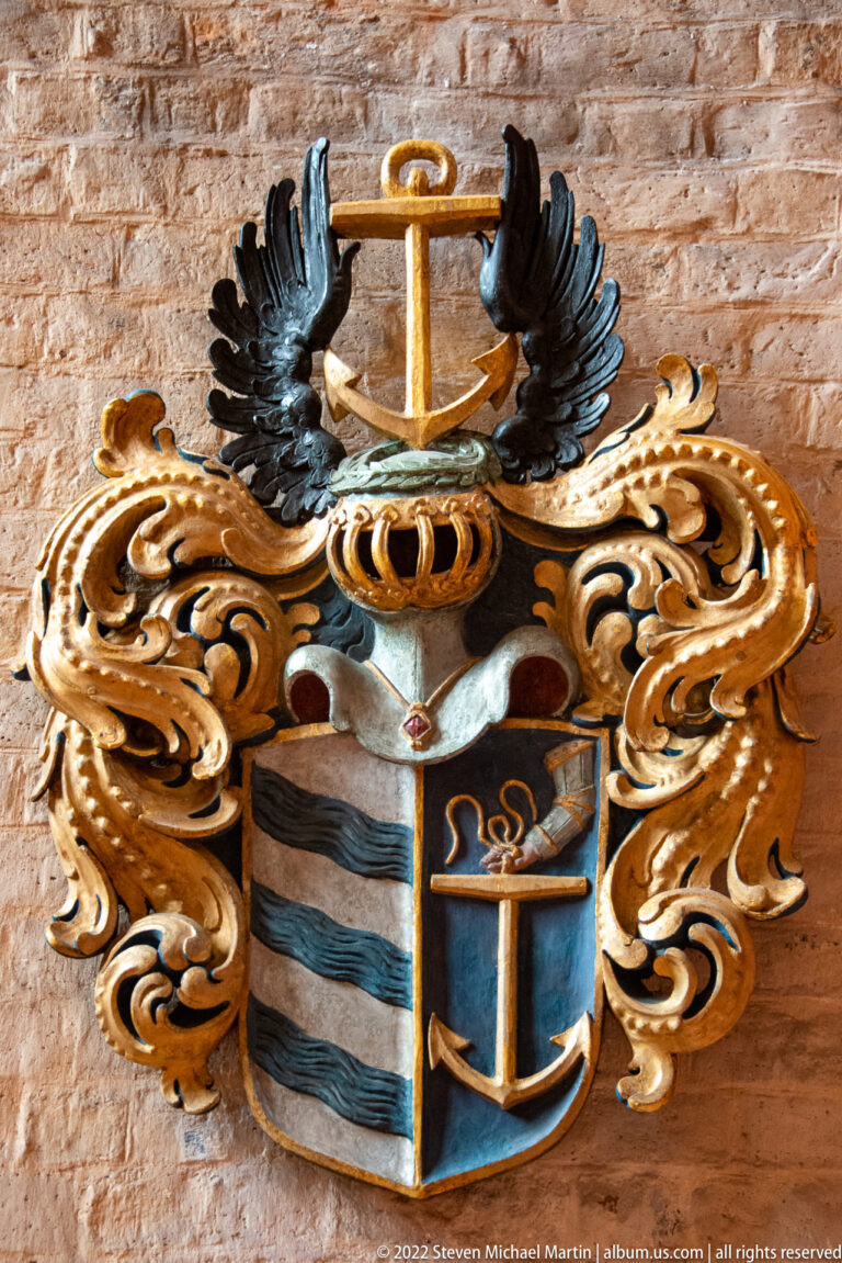 Gilded heraldic carving with quartered shield, anchor motifs, black wings, and scrolling foliage mounted on brick.