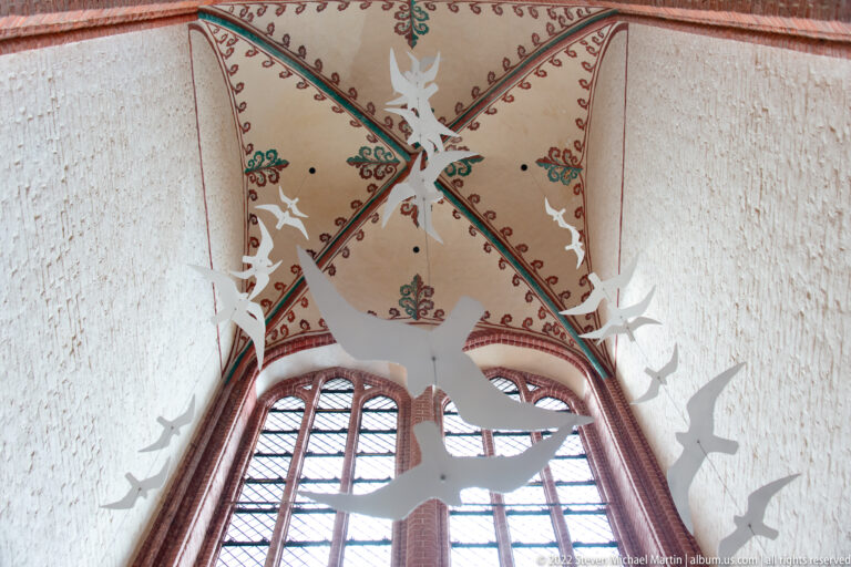 White bird silhouettes suspended beneath a ribbed church vault in front of a tall window.