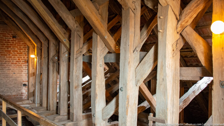 Interior of a historic roof with massive timber posts, beams, diagonal braces, and visible joinery beside brick walls.