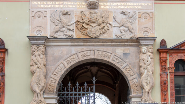 Ornate carved stone portal with heraldic crest, inscription, relief figures, and arched opening.