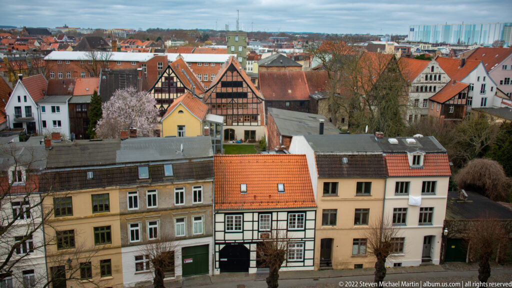 Elevated view across Wismar houses and red-tiled roofs toward distant harbor cranes and masts.