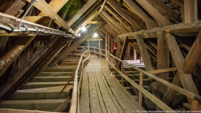 Wooden walkway with handrails passes through dense historic roof timbers under electric lights.