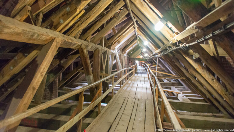 Long plank walkway recedes between steep rafters, braces, handrails, and lights inside a historic timber roof.
