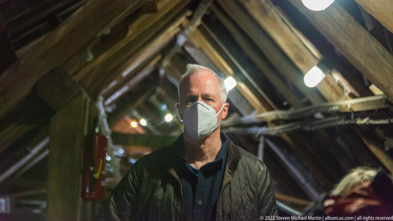 Masked visitor stands among heavy roof timbers, lights, and utility lines inside a narrow historic tower space.