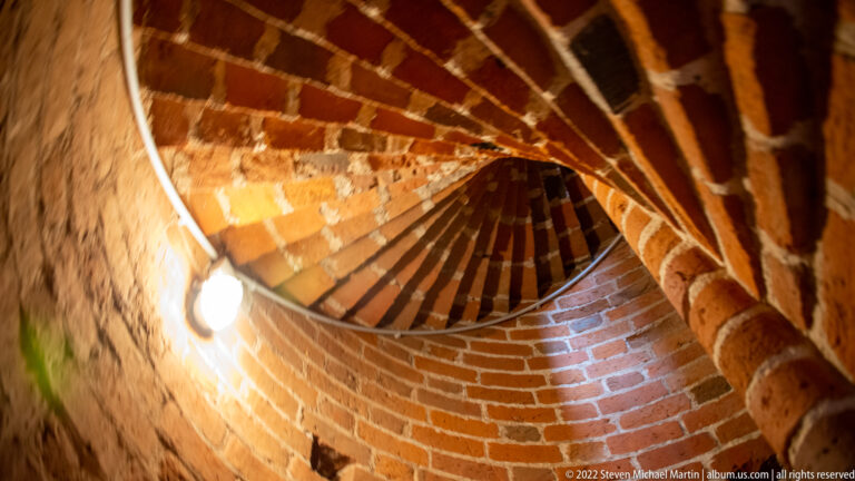 Narrow brick spiral stair curves upward through a cylindrical shaft lit by warm electric light.