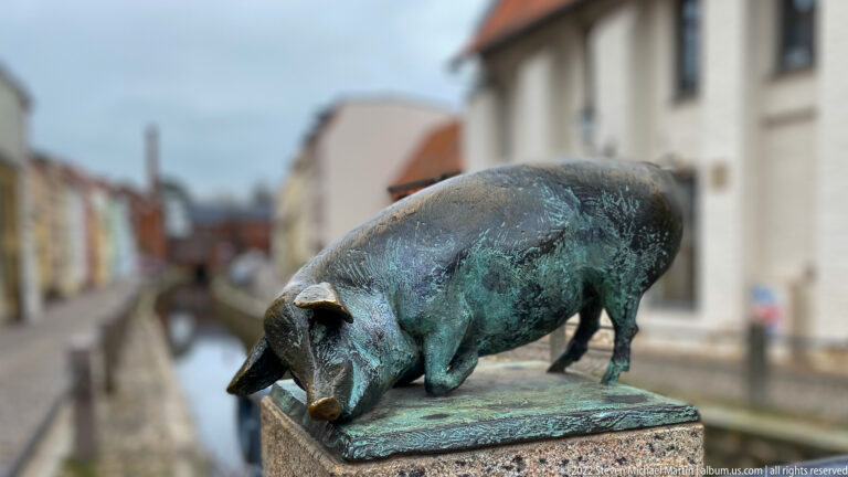 Green-patinated bronze pig stands with head lowered on a bridge post beside Wismar's canal.