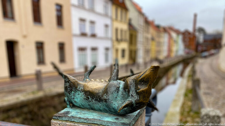 Bronze pig lies on its back with legs raised on a bridge post beside the canal.