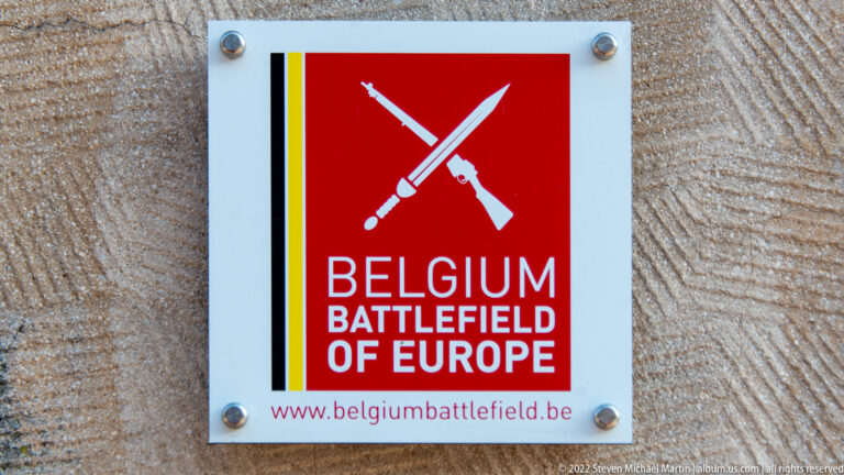 Red Belgium Battlefield of Europe sign with crossed weapons and Belgian tricolor stripe.