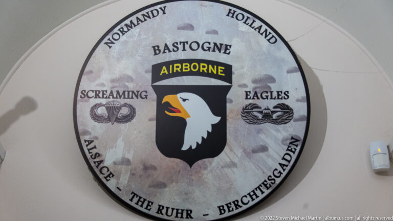 Circular wall emblem with Bastogne, airborne eagle, and campaign names including Normandy, Holland, Alsace, Ruhr, and Berchtesgaden.