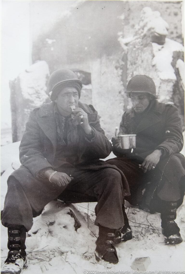 Black-and-white wartime photograph of two helmeted soldiers sitting in snow, one holding a metal cup.