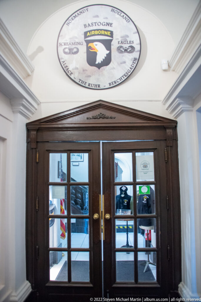 Dark wooden glass-paneled double doors beneath a circular Bastogne airborne emblem, with exhibits visible beyond.