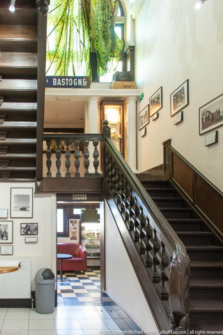 Dark wooden staircase with carved balustrade, framed photographs, and BASTOGNE sign above the upper doorway.