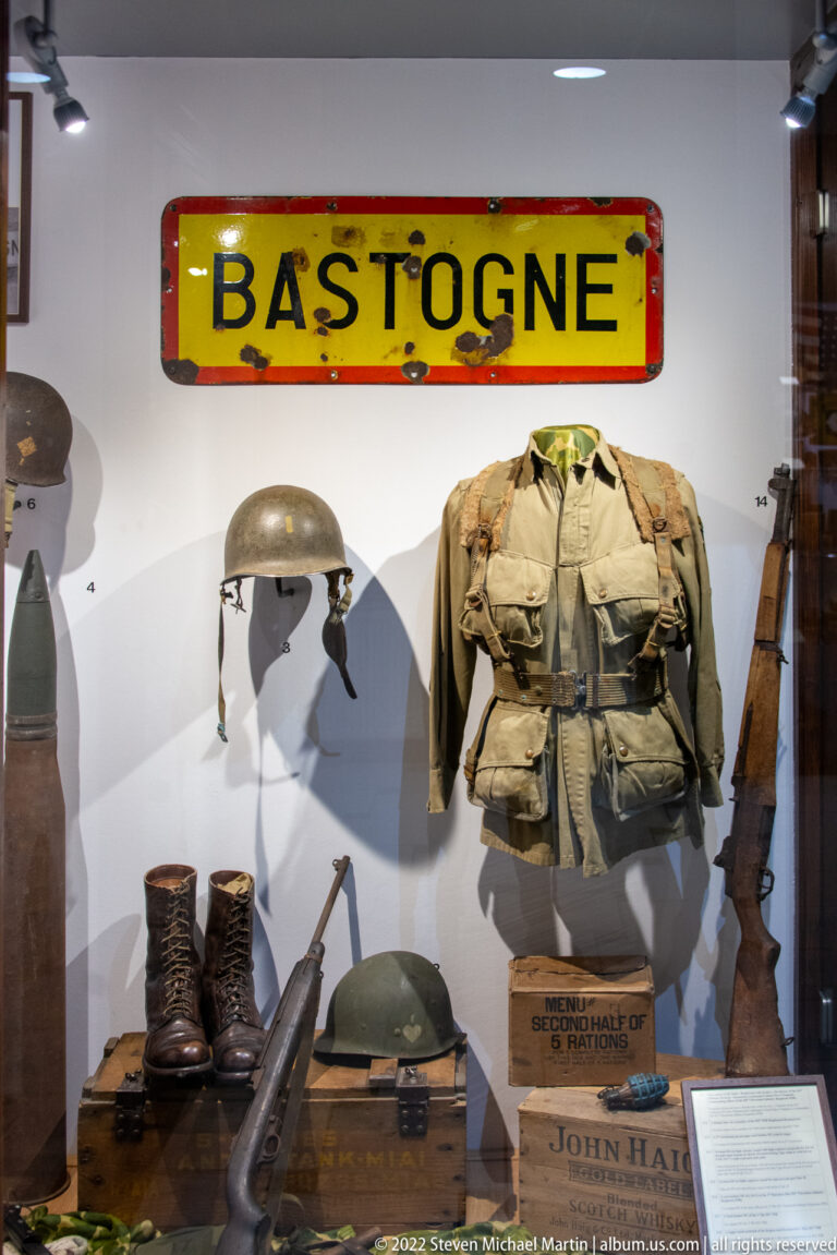 Museum case with BASTOGNE sign above a field jacket, helmet, boots, rifle, and assorted military gear.