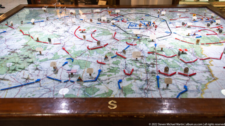 Glass-covered tabletop battle map with raised unit markers, colored arrows, pins, and labels.