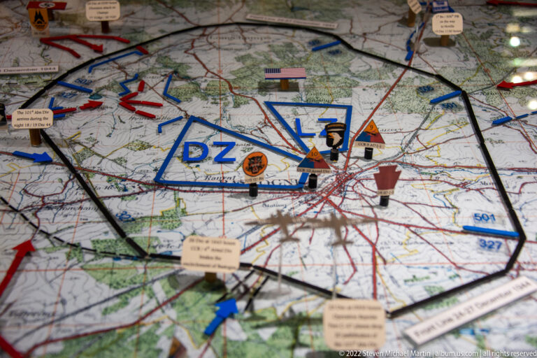Close-up of tabletop battle map with blue geometric markers, red and black lines, unit blocks, and labels around Bastogne.