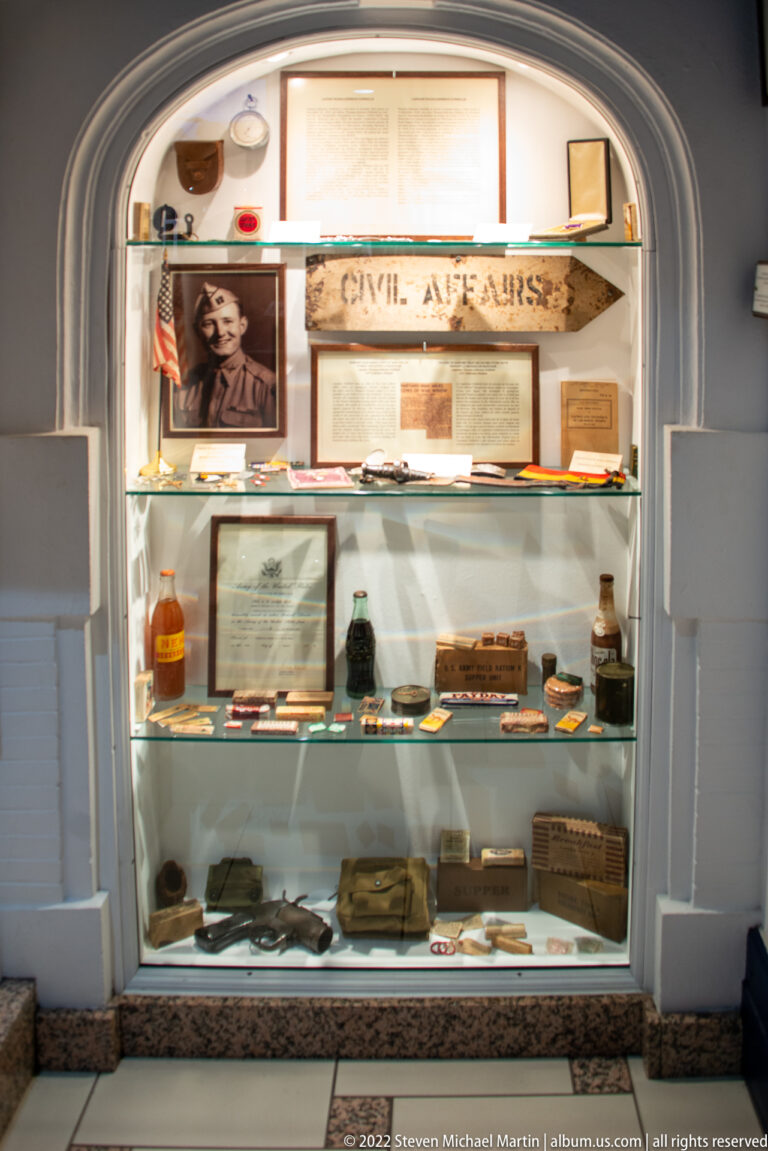 Recessed CIVIL AFFAIRS display case containing documents, photographs, bottles, pouches, and small wartime objects.