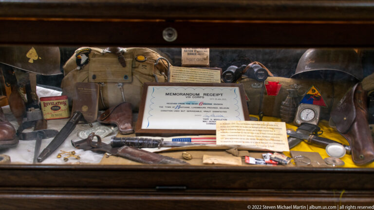 Museum case with helmet, canvas pack, goggles, memorandum receipt, bottles, tools, and assorted field gear.