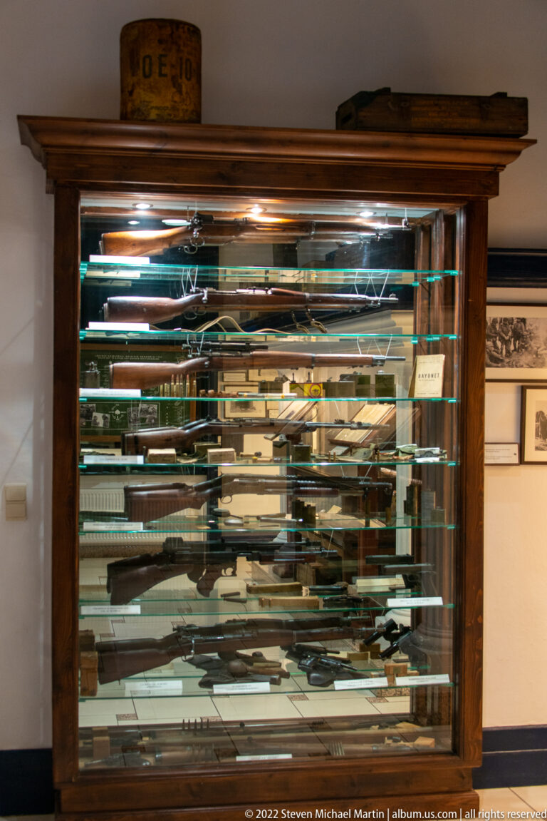 Tall glass-fronted wooden cabinet densely filled with rifles, long guns, and edged military weapons.