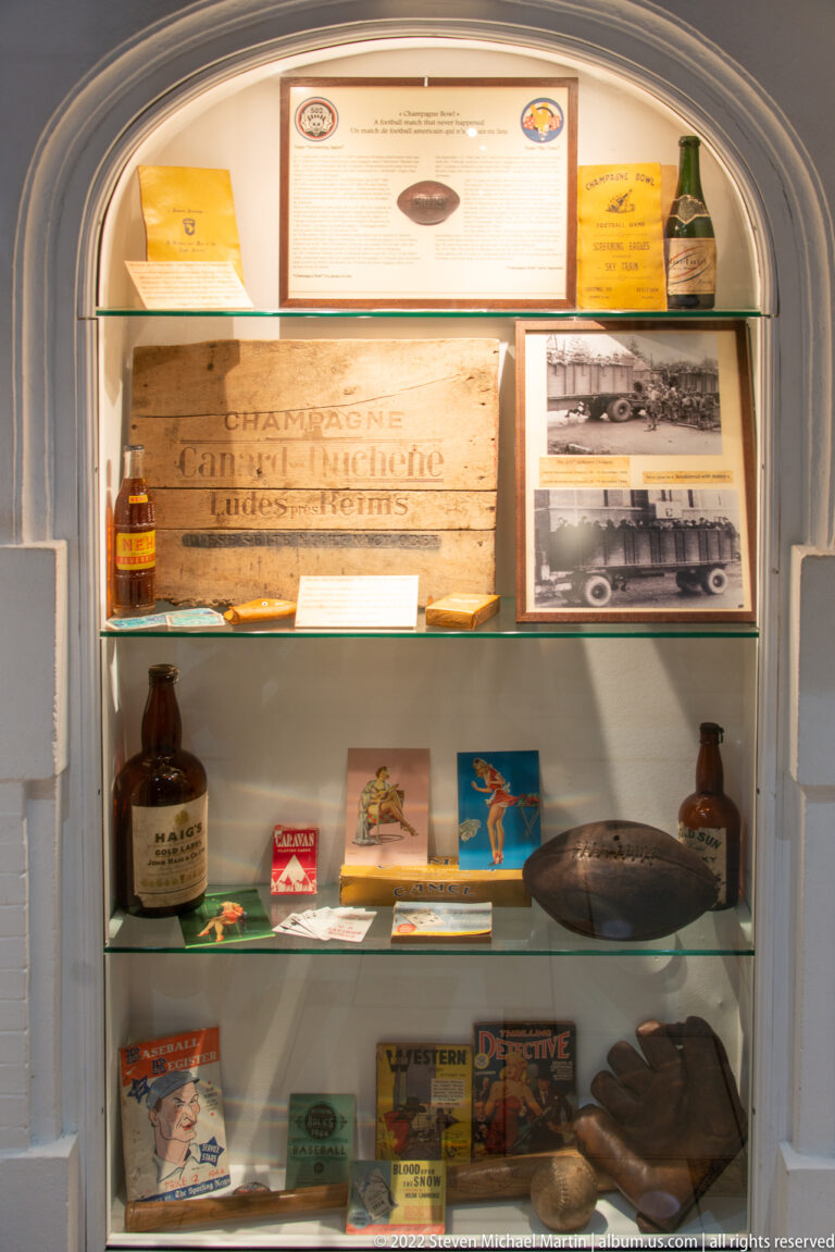 Glass display case with wartime books, bottles, photographs, shoes, helmet, and printed ephemera.