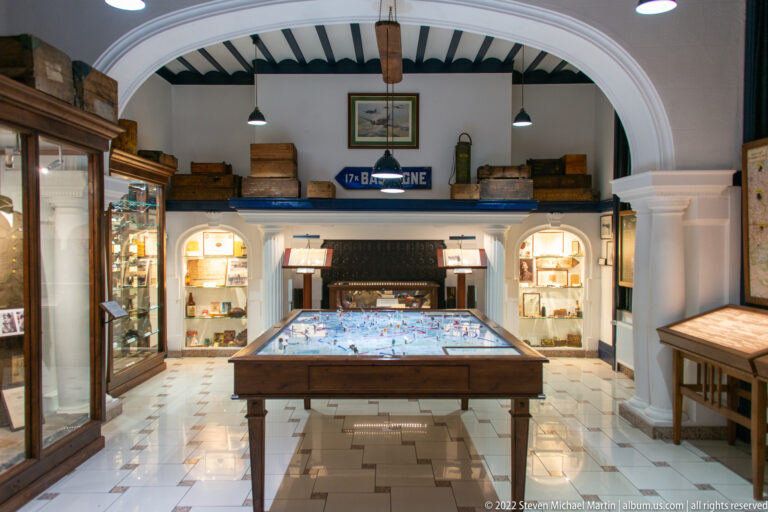 Central campaign map table in a museum gallery surrounded by illuminated artifact cases, cabinets, and period containers.