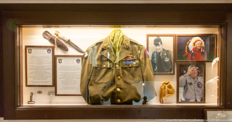 Decorated U.S. Army uniform displayed with portrait photographs, labels, and small personal objects in a museum case.