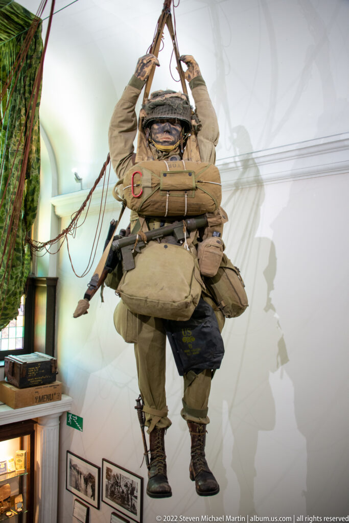 Suspended paratrooper mannequin wearing helmet, harness, webbing, and bulky field equipment beneath parachute lines.