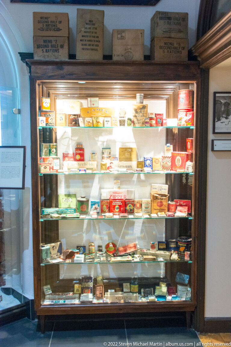 Large museum case densely filled with wartime cartons, tins, packets, and other small military consumables.