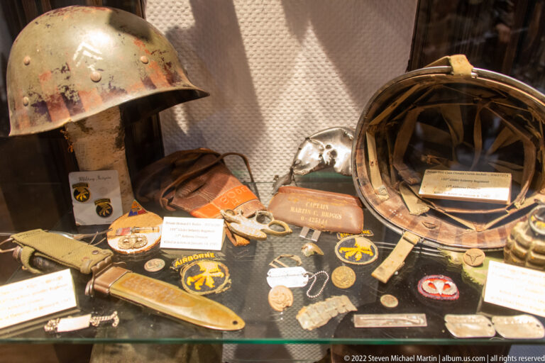 Close display of military helmets, cloth patches, badges, straps, medals, and other small personal artifacts.