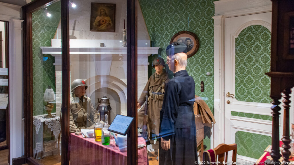 Reconstructed wartime room with uniformed figures, table, lamp, patterned wallpaper, and domestic furnishings behind glass.