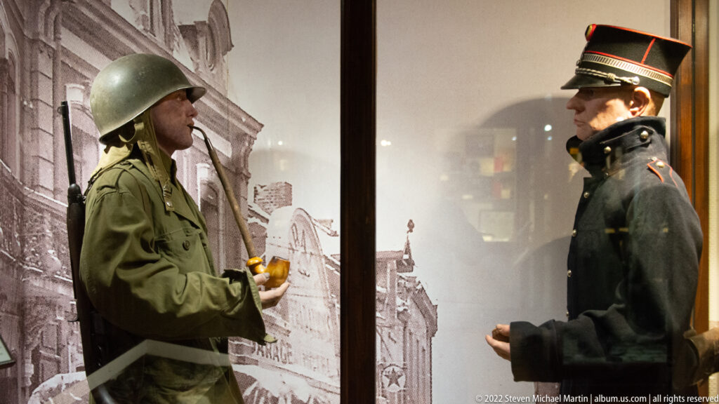 Helmeted U.S. soldier facing a dark-uniformed official with peaked cap against an urban streetscape backdrop.